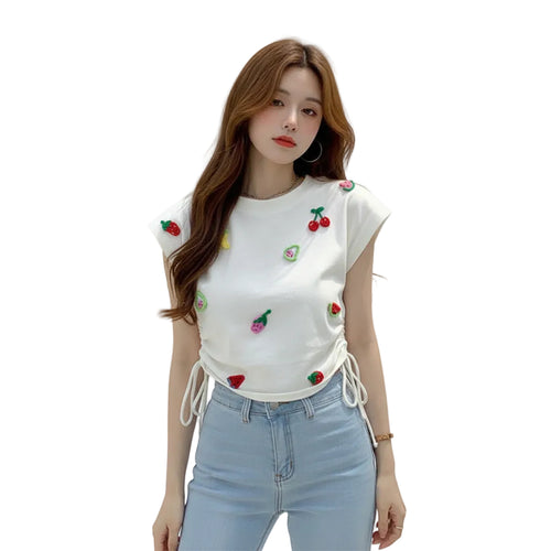 Woman wearing a white crop top with colorful fruit designs and light blue jeans, standing next to a car.