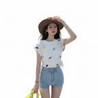 Woman in a white t-shirt with fruit patterns and denim shorts standing on a road with palm trees in the background.